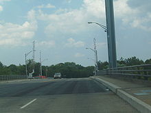A long bridge with a steel grate deck and lift towers gazing over cars and a pedestrian, who is on the side of the bridge jogging