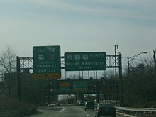 A mulitlane freeway in an urbanized area with two green signs over the road. The left sign reads exit 72 U.S. Route 9W Palisades Interstate Parkway Palisades Parkway Fort Lee exit upper left arrow only and the right sign reads Interstate 95 U.S. Route 1 U.S. Route 9 north George Washington Bridge