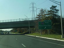 A six-lane freeway at an interchange with an overpass over the road and transmission lines running to the right. A green sign on the right side of the road reads exit 8A County Route 583 south Princeton Pike with an arrow pointing to the upper right