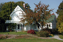 A white, two-story house with a one-story addition built at right angles to the main structure is surrounded by trees large and small, shrubs and other plants, and a lawn with gravel paths. The upper-floor windows are opaque and white, apparently boarded. Two chimneys protrude through a green shingled roof that appears to be in good condition.