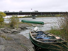Two canoes sit on the rocky bank of a river in front of a large concrete bridge with vehicles driving across it