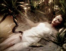 The body of a woman in a white dress floats on her back in a pond. Her body is submerged, and only her face, with open eyes, is above the water. There are plants near her, and a black snake swims through the water, its head over the top of her exposed thigh.