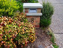 A squat, square red brick detached mailbox about three feet or 90 centimetres in height. Below a thin metal letter slot is the number 8.