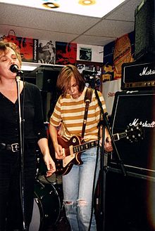 Linda Hopper stands behind a microphone singing with Ruthie Morris to her left playing guitar in front of a stack of Marshall Amps