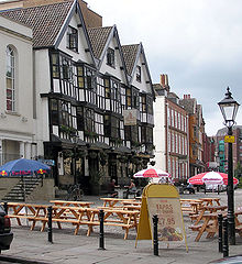 A seventeenth century timber framed building with three gables and a traditional inn sign showing a picture of a sailing barge. Some drinkers sit at benches outside on a cobbled street. Other old buildings are further down the street and in the background part of a modern office building can be seen.