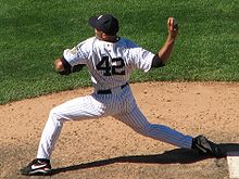 Mariano Rivera in a white pinstriped baseball uniform and navy blue cap stands on a dirt mound. He is striding forward to the left as he clutches a baseball behind his head. The back of his uniform reads "42" in navy blue numbers.