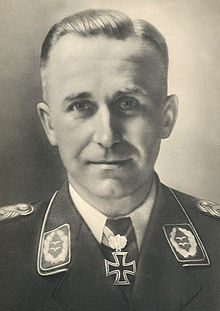 The head and shoulders of a man, shown from the front. He wears a military uniform, a white shirt with an Iron Cross displayed at the front of his shirt collar. His hair appears blond and short and combed back, his facial expression is a determined and confident smile; his eyes are looking into the camera.