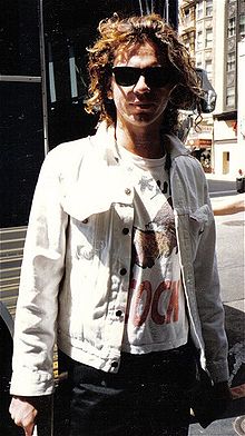 Upper body of a 26 year old man. He is standing with his arms alongside. He has collar length brown hair. His light coloured jacket is opened to reveal a white T-shirt with an obscured image and printing. He wears dark glasses and the left side of his face is partly in shadow. Over his right shoulder is the entry way to a building, with other buildings nearby.