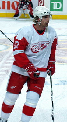 A Caucasian ice hockey player in his thirties. He is skating relaxed on the ice while looking to his right. He wears a white and red jersey, along with a white visored helmet.