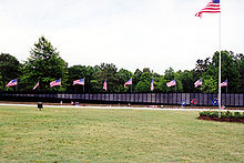 A green lawn with a wall in the background having several United States flags flying in the background and one tall flag pole with a high flying flag.