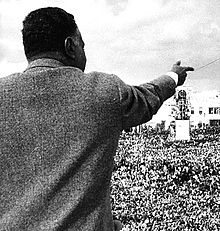 The back of a man in a gray jacket pointing forward, with a large crowd in front of him.