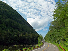 A two-lane road winding between two steep and wooded slopes. There is a small body of water on the left