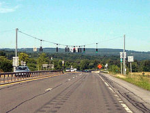 A view of the four-lane NY 5S as it approaches a traffic signal. NY 28 is accessed by turning left at the signal. In the background and distance are tree-covered mountains.