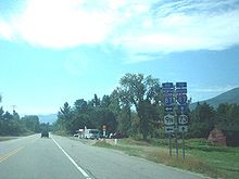 A two-lane highway in a wooded, mountainous rural area. To the right of the highway is a sign assembly instructing drivers to turn left for NY 9N and continue straight for NY 73.