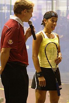 A man in a red shirt holds a microphone while a young woman in a yellow shirt holding a racket sticks her tongue out.