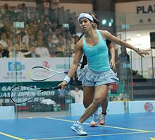 An indoor squash-court; a female player in light blue sportswear, a short pleated skirt, balancing for a shot, facing the camera and mostly obscuring her partner who is behind her. Both their rackets are at knee level on their left. A crowd is in the background, behind glass.