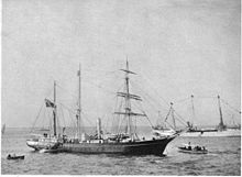  A three-masted ship with sails furled, short funnel amidships, flag flying from the stern on left of picture. Two small boats are close by, and a larger vessel decked with bunting is visible in the background.