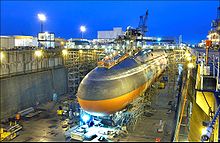 Black submarine with orange paint from cheatline down in drydock at nightfall.