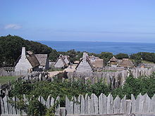 A modern day photograph of a village consisting of small, primitive wooden houses.  Most of the houses have thatched roofs.  In the distance is a large expanse of ocean and a clear blue sky.  The village is surrounded by a wall consisting of tall, thick wooden planks.