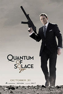 A man in a business suit holding a machine gun stands on a desert. To his left is the title "Quantum Of Solace" in black letters – except the 'O's, which are golden and make a diagonal straight line with a 7 resembling a gun.