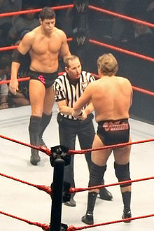 Two men face off in a wrestling ring with a referee between them. The one facing the camera has short black hair, and is wearing short black wrestling tights with the letters 'CR' upon them in pink with black boots and a black elbow pad on his right arm. The man facing away from the camera has longer, light brown hair and the word 'villain' is written upon the back of his short black wrestling tights.