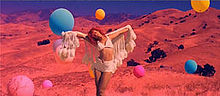 Rihanna standing in the middle of a field, with a shawl, surrounded by floating balloons