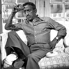 Black-and-white photograph of an elderly African-American man wearing a striped shirt, grey trousers, a watch and various jewelry, sitting hunched on a sofa with a sombre expression.