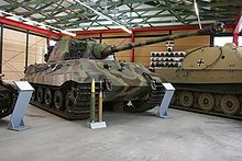 A three quarters view of a large tank with a flat-faced turret, dull yellow, green and brown wavy camouflage, on display inside a museum. The frontal armor is sloped. The long gun overhangs the bow by several meters. Two waist-high cartridges sit on their bases infront of it.