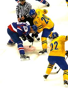 A faceoff during an ice hockey game. The player at the top left has directed the puck to his teammate next to him as his opponent, bent over, looks on.