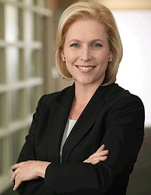 A portrait shot of a smiling, middle-aged Caucasian female (Kirsten Gillibrand) looking straight ahead. She has long blonde hair, and is wearing a dark blazer with a grey top; on her left lapel is a gold pin that reads "United States Senator". She is placed in front of a dark background.