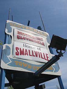 A white billboard with blue ribbon painted around the edges. In the ribbon, the words "Cloverdale Town Centre" appear in yellow surrounding two bannders. The top banner reads, "Destination Cloverdale", while the bottom reads "Home of Smallville".