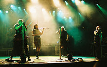 Four singers perform in the spotlights on stage. One is a female in  shorts, a gauzy beige skirt and a leather belt, and plays the fiddle,  The males are wearing black sleeveless shirts, black pants and have military-looking gear. A rock band is in the background.