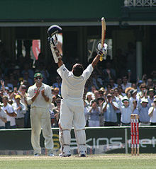 A man wearing cricket whites and batting pads, standing next to a wicket with his arms aloft. He is holding a helmet in his left hand, and a cricket bat in his right hand. In the distance, supporters are applauding.