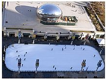 An ice skating rink with a few skaters and a large metallic sculpture in the background