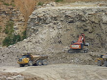 A photograph showing quarrying operations with a mechanical digger in the centre extracting rock from a cliff face. To the left a lorry is just leaving with a full load of rock and to the right another lorry is waiting to collect its load from the digger.
