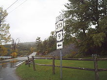 A two-lane highway in a forested area on a rainy day. A sign assembly in the foreground indicates that NY 373 and US 9 can be accessed by following the aforementioned two-lane highway.