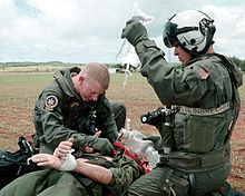 Color photograph of a United States Navy hospital corpsman listening for correct placement of an endotracheal tube in a simulated trauma victim during a search and rescue exercise. His assistant is holding a bag of intravenous fluid.