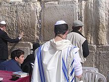 A man wearing a white robe with blue stripes stands in front of a stone wall.