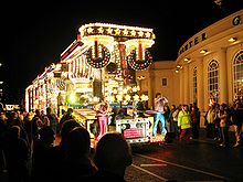 A large vehicle lit by multi-coloured lamps and carrying people in brightly-coloured coustumes in front of a large crowd on a darkened street.