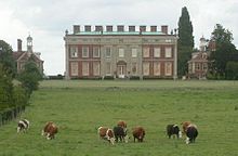 Large two-storey brick building, flanked by two smaller brick pavilions. In front of the larger building is a herd of cattle.