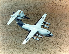 Top view of cargo aircraft in-flight, trailed by a fighter chase plane. Under each un-swept wing are two engines suspended forward ahead the leading edge.