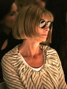 A woman wearing sunglasses and a gray-and-white striped top in a dark background looking to the right