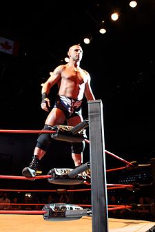 A male with black taped hands wearing black trunks, knee pads, and boots standing on the ropes in a wrestling ring corner.