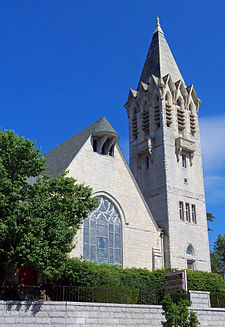 A bright-colored stone building with a steep pointed roof and a tall square pointed tower on its right. It has a large stained-glass window in front and some decorative touches.