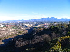 A small stream winds past in the lower left foreground while residential subdivisions sprawl over the hills on both sides. Mountains rise in the distance beneath an empty, pale blue sky.