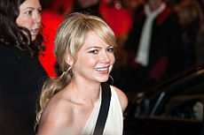 An image of a smiling Caucasian women. She has her blonde hair in a ponytail and is wearing a one shouldered white dress with a black horizontal stripe.