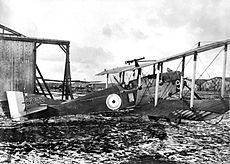 Side view of military biplane with pilot in cockpit, parked on landing ground