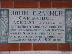 A tablet formed of six standard sized tiles, bordered by green flowers in the style of the Arts and Crafts movement. The tablet reads "John Cranmer Cambridge aged 23, a clerk in the London County Council who was drowned near Ostend whilst saving the life of a stranger and foreigner, August 8, 1901".