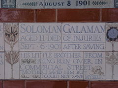 A tablet formed of five tiles of varying sizes, bordered by yellow and blue flowers in an art nouveau style. The tablet reads "Solomon Galaman, Aged 11 died of injuries, Sept 6 1901 after saving his little brother from being run over in Commercial Street, 'Mother I saved him but I could not save myself.'"