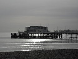The end of a pier under cloudy skies, illuminated by sunlight reflecting off a calm sea.  There is a small stretch of rocky beach in the foreground.